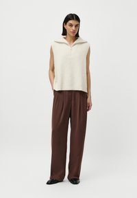 Femeie îmbrăcată cu un vestă tricotată crem cu fermoar, fără mâneci, pantaloni maro largi și încălțăminte neagră, stând în fața unui fundal alb simplu.