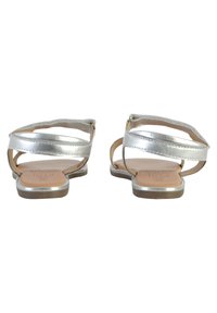 Sandales plates argentées avec des brides croisées, présentant une finition métallique lisse et une semelle intérieure beige. Design ouvert à l'arrière avec une semelle minimaliste.