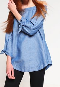 Modrá šambráová off-the-shoulder blůza s dlouhými rukávy, která má na manžetách mašličkové detaily. Volný střih a zakřivený lem.