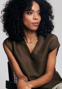 Brązowa bluzka krótkim rękawem z dekoltem w serek, gładka w dotyku. Złoty naszyjnik podkreśla dekolt. Modelka siedzi z kręconymi włosami.