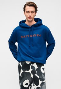 Μπλε μπλούζα με κουκούλα από μείγμα βαμβακιού, με λογότυπο εμπρός σε πορτοκαλί γράμματα. Συνδυάζεται με ασπρόμαυρο παντελόνι με σχέδιο.