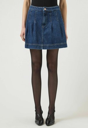 Kvinde iført en blå denim mininederdel, sorte transparente strømpebukser og sorte højhælede sko, stående foran en ensfarvet baggrund.