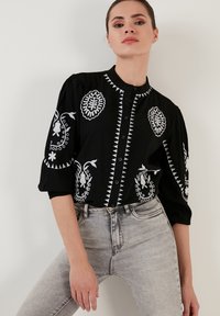 Schwarzes Hemd mit Knöpfen und weißer Stickerei, das florale und geometrische Muster aufweist, Dreiviertelärmel hat und einen runden Halsausschnitt besitzt.