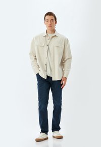 Camisa de botón de color beige claro con dos bolsillos en el pecho, combinada con jeans azul oscuro y zapatillas blancas con suelas marrón claro. Diseño sencillo, ajuste casual.