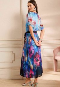 Hosszú, pliszírozott maxi ruha kék színben, rózsaszín és lila virágmintával. Átlátszó, rövid ujjai és fekete öve hangsúlyozza a derekát. Ezüst magassarkú cipők egészítik ki a megjelenést.
