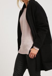 Svart, lång kappa i texturerad tyg, kombinerad med en ljusrosa stickad tröja och svarta läderbyxor; har framfickor.