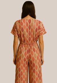 Jumpsuit à motifs rouge et beige avec un design de feuillage, manches courtes et taille nouée. Le tissu texturé rehausse le détail visuel.