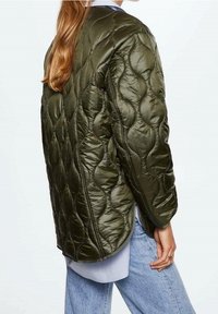 Zelená prošívaná bunda s hladkým povrchem, zaoblenými tvary a uvolněným střihem. Nositá přes světle modrou košili s volnými džíny.