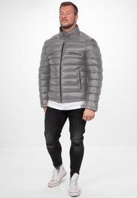 Grijze pufferjack met een hoge kraag, met horizontaal gestikte secties, twee ritszakken op de borst en een logo op de schouder.