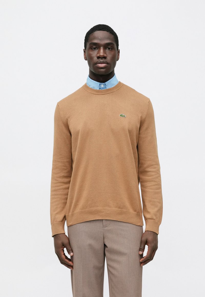 Rjavi pleteni pulover z okrolim ovratnikom in zelenim logotipom Lacoste. Nošen čez svetlo modro srajco z ovratnikom in kombiniran s bež hlačami.