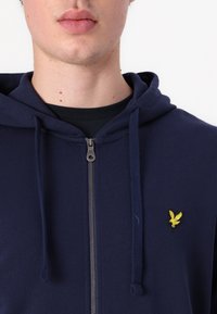Tamnoplava zip-up dukserica napravljena od mekog materijala, s malim žutim vezom ispogo orla na prsima i podešavajućim špagama na kapuljači.
