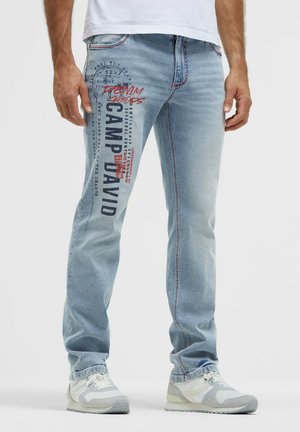 Mann trägt hellblaue Jeans mit rotem und schwarzem Text "Camp David" auf dem linken Oberschenkel, weiße Sneaker und ein weißes Hemd.
