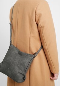 Borsa a tracolla in pelle grigia con una finitura testurizzata, caratterizzata da una singola tracolla regolabile e accessori in argento, indossata sopra un cappotto beige.