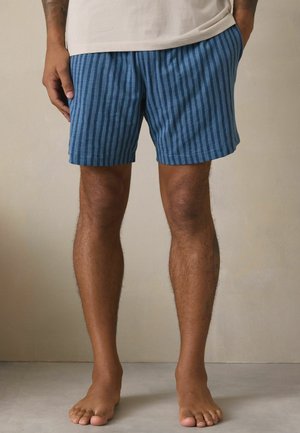 Mees seisab paljajalu, kandes sinise triibulise lühikese püksid ja beeži särki, käed taskutes, tühja tausta ees.