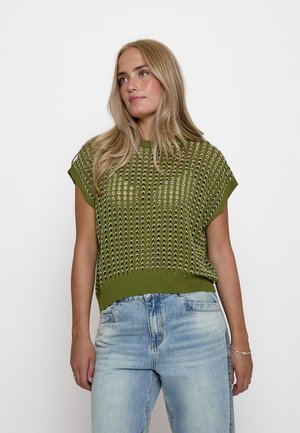 Donna con lunghi capelli biondi che indossa un top a maglia verde a maniche corte con motivi traforati e jeans chiari, in piedi davanti a uno sfondo bianco.