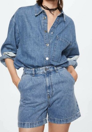 Chemise en denim bleu clair avec un devant à boutons, des manches retroussées et une poche. Assortie à un short taille haute en denim, également bleu clair.
