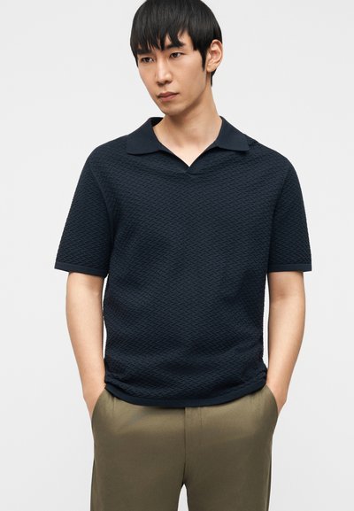 Νέος άνδρας με ίσια μαύρα μαλλιά, που φοράει μπλε σκούρο πόλο μπλουζάκι με κοντά μανίκια και υφή, καθώς και χακί παντελόνι, με τα χέρια στις τσέπες.