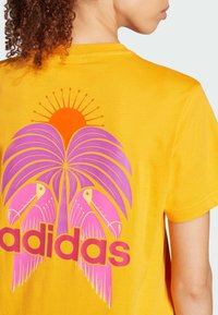 Žluté bavlněné tričko s grafickým potiskem palmy a dvou tukánů v růžové a oranžové barvě, s nápisem "adidas" v tučném růžovém písmu.