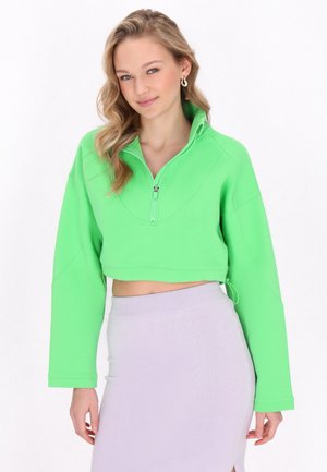Neon grøn cropped sweatshirt med højt krave og lynlås foran. Har lange, brede ærmer og en rundet kant. Sammen med et lyse-lilla nederdel.