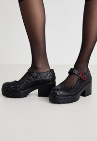 Svarta glittriga Mary Jane-skor med en tjock klack, som har en utskärningsdesign och en kontrasterande röd detalj på remmen. Texturerad ovandel, stabil sula.