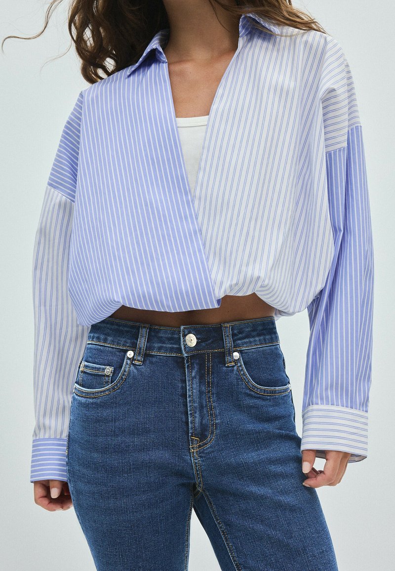 Femme portant un jean bleu taille haute et un chemisier cache-cœur rayé bleu et blanc par-dessus un débardeur blanc, visible du cou aux hanches.