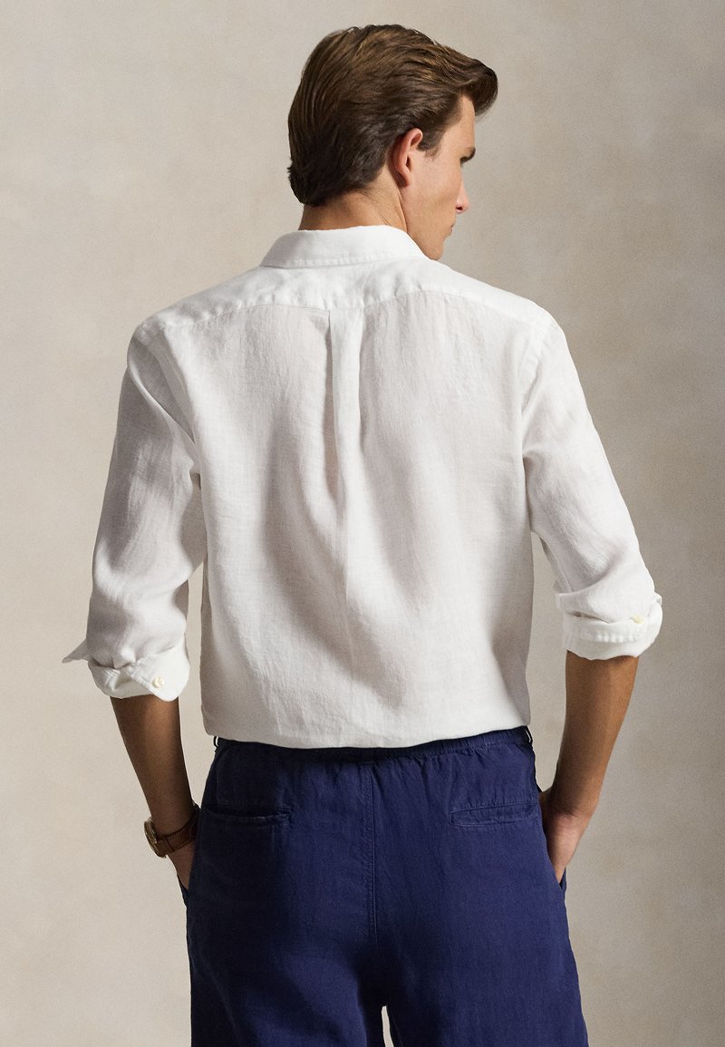 Mann trägt ein langärmeliges weißes Hemd mit hochgekrempelten Manschetten und dunkelblaue Hose, steht mit den Händen in den Taschen und blickt von der Kamera weg.