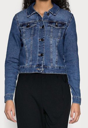 Vrouw draagt een blauwe spijkerjas met knoopsluiting, borstzakken en lange mouwen, gecombineerd met een zwarte broek.