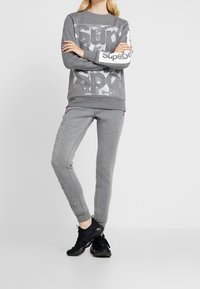 Felpa grigia con stampa grafica, abbinata a pantaloni jogger grigi a sigaretta con dettagli del logo. Indossata con scarpe da ginnastica nere.