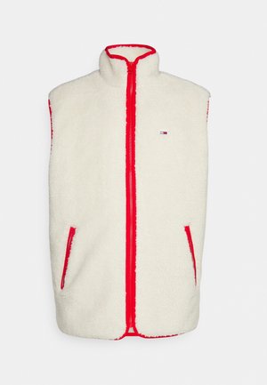 Roomkleurig fleecevest over een rood hoodie, gecombineerd met witte gestructureerde broek en zwarte sneakers. Voorzien van een ritssluiting en contrasterende bies.