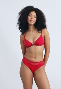 Rød lingeri-set bestående af en blød BH med tynde stropper og matchende gennemsigtige hipstertrusser. Begge dele har en glat tekstur med blonderdetaljer.