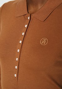 Brązowa koszulka polo z fakturowanej tkaniny, z kołnierzykiem i siedmioma białymi guzikami. Haftowane logo w złotym kolorze na lewej piersi.