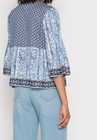 Blusa blu e bianca con motivo, caratterizzata da maniche a volant e uno sprone a contrasto in blu navy. Il tessuto appare leggero con dettagli floreali.
