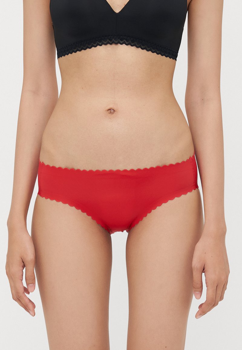 Red bikini briefs made of smooth fabric with a scalloped waistband and leg openings, featuring a snug fit. Black top visible above.