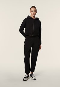 Haut de survêtement noir court avec fermeture éclair, assorti à un pantalon fuselé noir ; porté avec des baskets noir et blanc. Texture de tissu lisse.