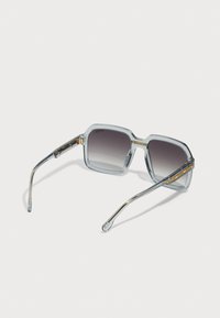 Occhiali da sole esagonali chunky con montatura trasparente blu chiaro, lenti grigie sfumate e dettagli metallici dorati sulle aste.