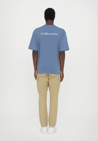 T-shirt en coton bleu à manches courtes, arborant un logo blanc "Polo Ralph Lauren" au dos. Associé à un pantalon cargo beige et des baskets blanches.
