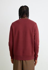 Bordeaux sweatshirt met ronde hals, ribbelmanchetten en -zoom. Zachte textuur, ontspannen pasvorm, gecombineerd met bruine geruite broek met een roestpatroon.