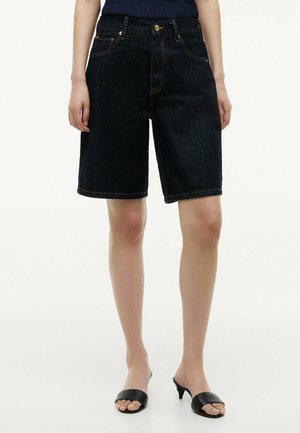 Kvinde iført mørke denim knælange shorts og sorte åbne sandaler med hæl, stående mod en lys baggrund.