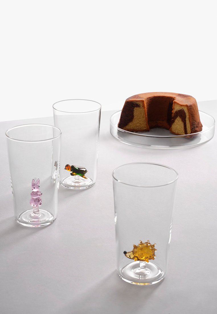 Przezroczyste szklanki z kolorowymi dekoracyjnymi figurkami (królik, ptak, jeż) w środku, obok okrągłego ciasta marmurkowego na przezroczystym talerzu.