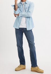 Ljusblå denimskjorta med spetsig krage, lagd över en vit t-shirt. Mörkblå jeans och bruna mockaskor, visade i en avslappnad ställning.