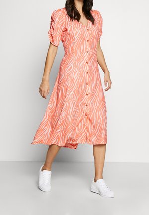 Femme portant une robe midi corail et blanche à imprimé zébré, manches bouffantes et boutons sur le devant, assortie de baskets blanches, debout contre un mur blanc.