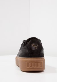 Svarta mockaskor med en tjock brun gummisula. Har en guldfärgad Puma-logotyp och strukturerade sidostråk på en delvis vadderad tunga.