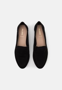 Mocassins en suède noir avec une texture lisse, un bout arrondi et des accents de couture. La semelle intérieure arbore le nom de la marque en lettres dorées.
