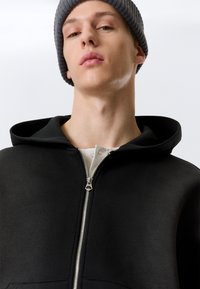 Sweat à capuche noir avec fermeture éclair, portant une chemise blanche boutonnée en dessous, et un bonnet tricoté gris. Texture de tissu lisse.