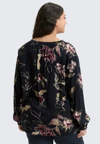 Frau mit langen, dunklen Haaren, die eine lockere schwarze Bluse mit pinken und grünen Blumenmustern trägt, mit dem Rücken zu einem schlichten, hellen Hintergrund.