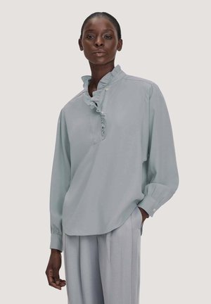Vrouw draagt een lichtblauwe blouse met geplooide kraag en lange mouwen, gecombineerd met loszittende lichtgrijze broek, staand tegen een effen achtergrond.