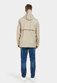 Beige vattentät jacka med hög krage, ryggslå och långa ärmar. Bärs över blå jeans och vita skor. Slät textur.