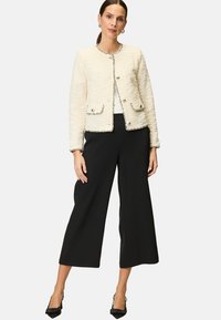 Ecru gewobene Jacke mit rundem Ausschnitt, kontrastierenden Verzierungen, zwei Fronttaschen und metallischen Knöpfen, kombiniert mit schwarzen weiten Hosen.