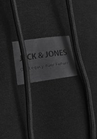 Sudadera negra con una textura suave que presenta una etiqueta rectangular negra grabada con "JACK & JONES" y "Nuestro Legado Tu Futuro."