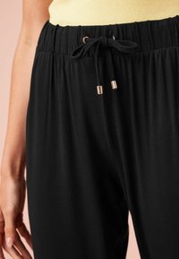 Pantaloni neri con vita elastica e cordoncino con estremità in metallo dorato; texture morbida, taglio rilassato e senza motivi visibili.